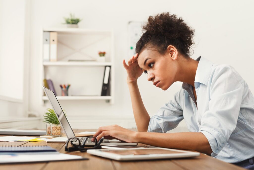 Woman with medium-dark skin tone hunches over laptop with strained facial expression.