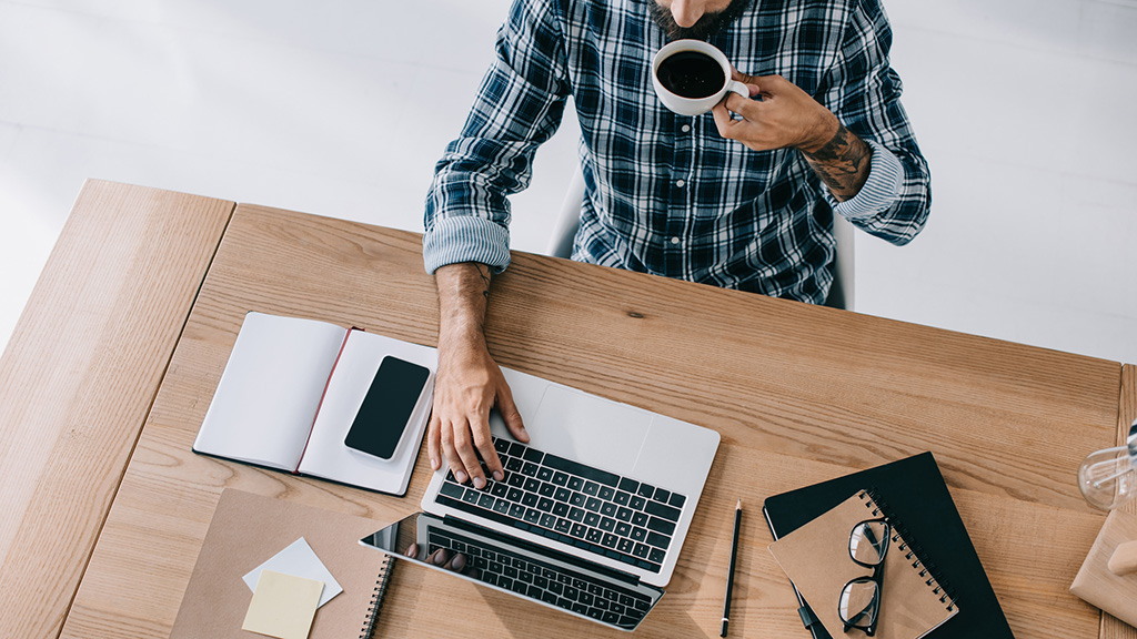 Overhead view of middle-aged man sipping coffee while typing on laptop at desk.