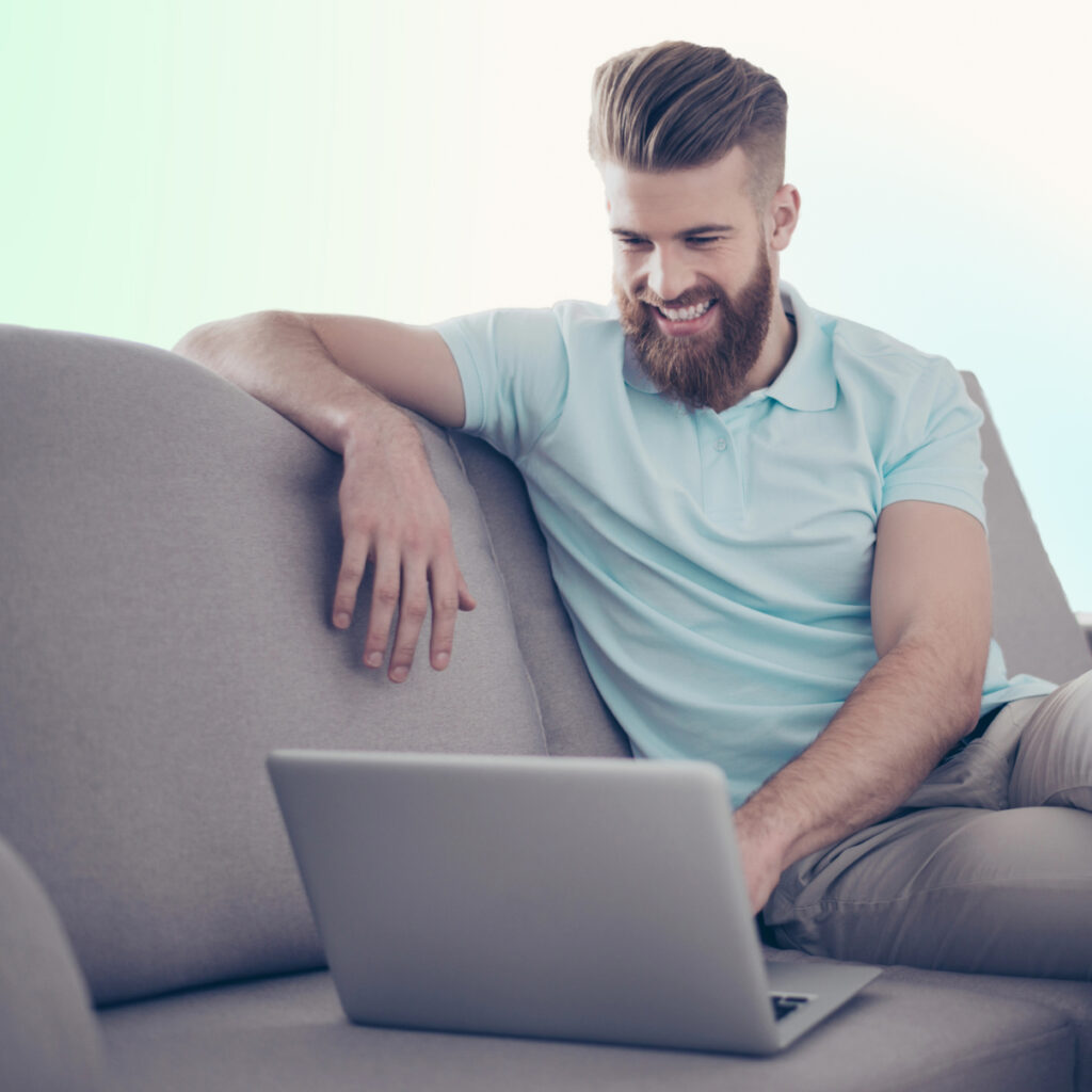 Bearded man with light brown hair smiling at laptop on couch.