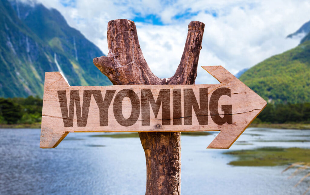 Arrow-shaped wooden sign reading 'Wyoming,' set against scenic mountains, lake, and cloudy blue sky.